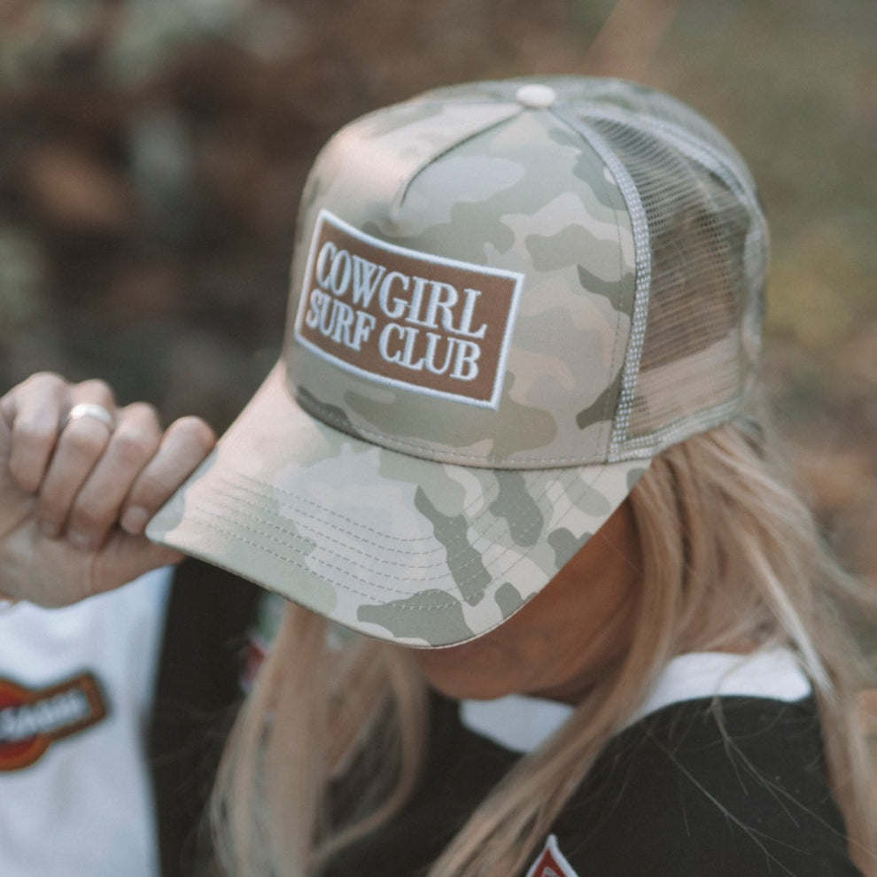 Person wearing a camouflage cap with 'Cowgirl Surf Club' logo outdoors.