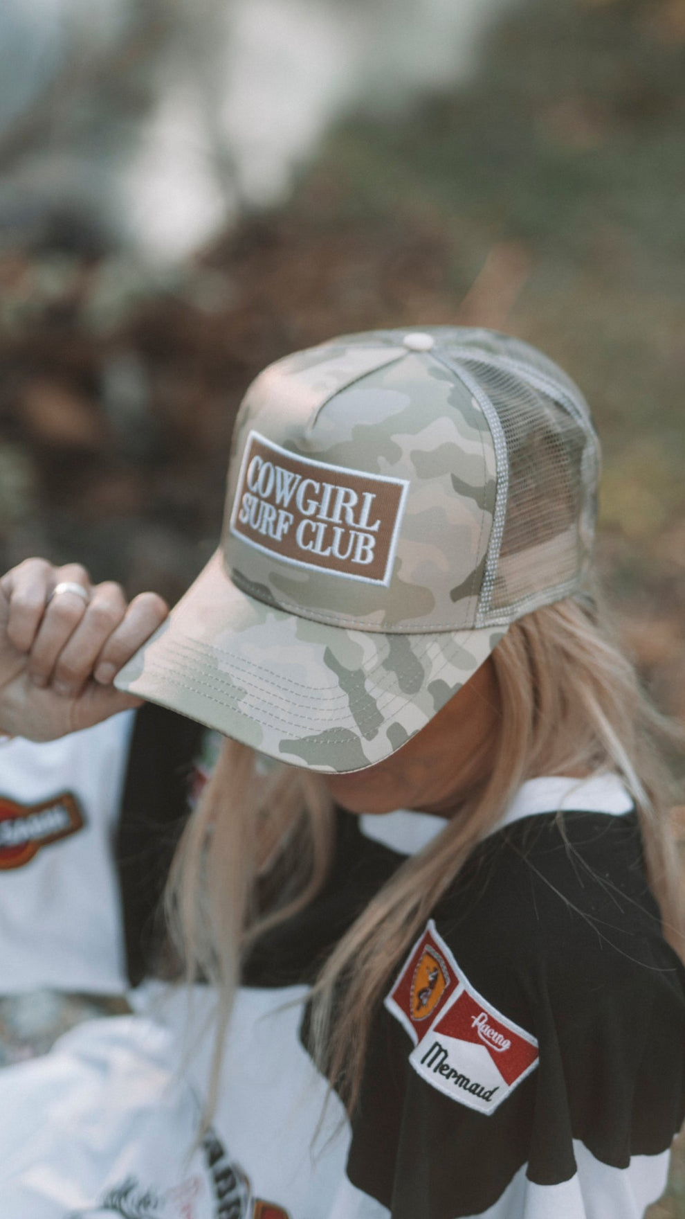 Person wearing a camouflage cap with 'Cowgirl Surf Club' logo outdoors.
